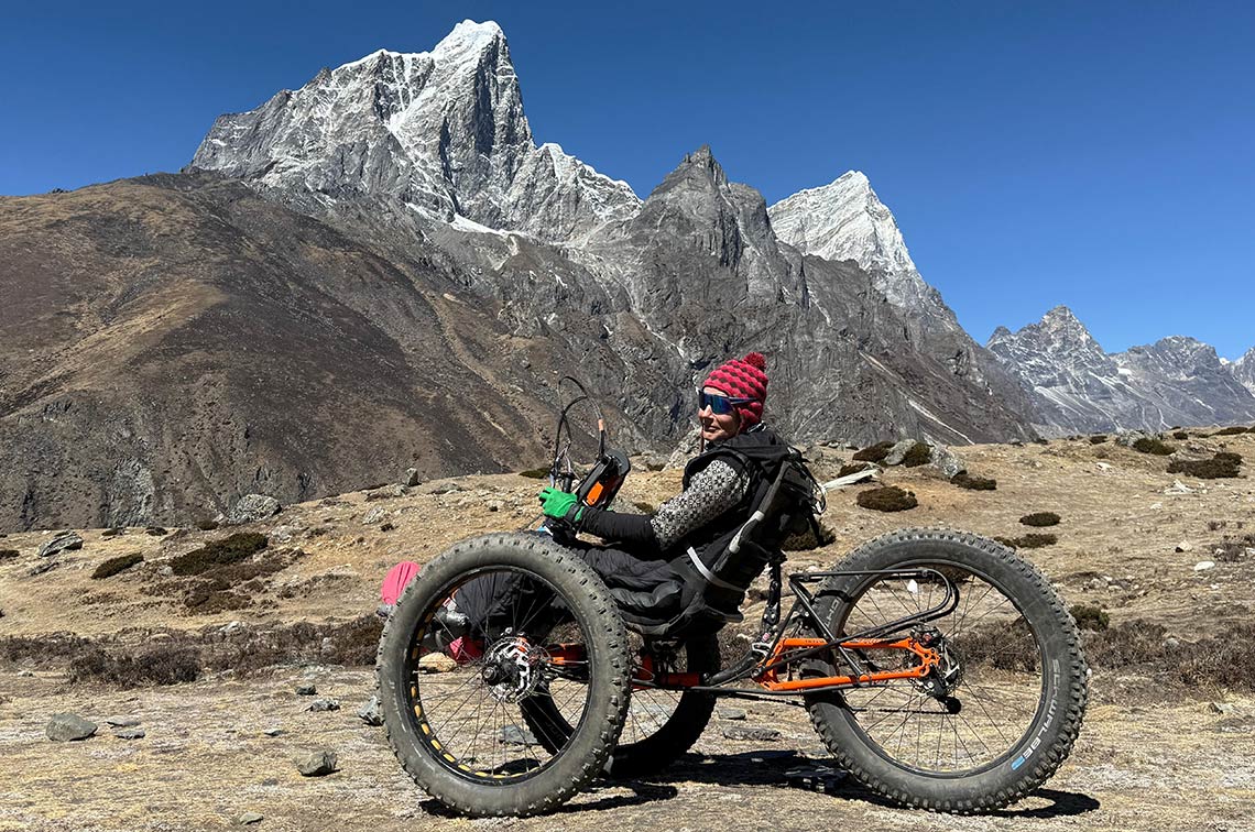 Adventurer Karen Darke sits semi-reclined in an all-terrain hand bike with three large, chunky tyres and an orange chassis on a rocky path in front of several towering, snow-topped Himalayan mountains and a clear blue sky. There are small patches of grass among the rocks. She�s wearing a red bobble hat with wrap-around sunglasses, pink boots, green gloves, a black and white patterned jumper and warm black trousers.