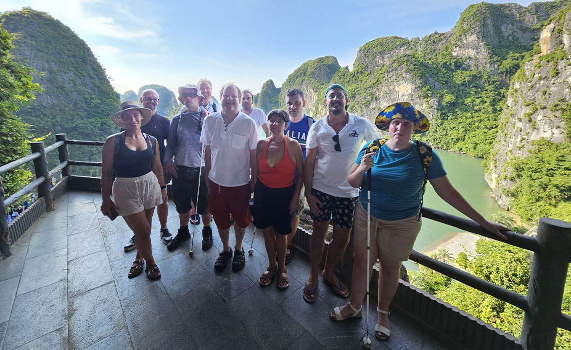 Rachel Nafzger wearing a blue stripy tshirt, blue floppy hat with yellow sunflowers, khaki shorts, sandals and a blue rucksack with a sunflower print and carrying a white cane is standing with a group of nine other blind travellers and guides on a balcony view point high up above Ha Long Bay in Vietnam. Behind them it�s sunny, with dramatic green-topped cliffs rising vertically out of the turquoise water far below.