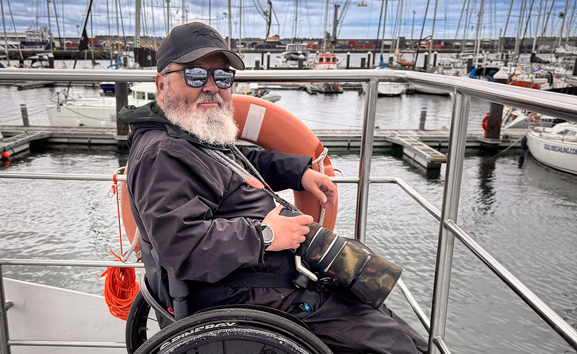 A close-up shot of Paul who has a grey beard, sitting in his wheelchair, which is pushed up against the railings on the top deck of a boat ready to go whale watching in the Azores. He is leaning one arm on the railings on the boat and wearing a cap and sunglasses despite a cloudy sky - and he is carrying a camera with a large telephoto lens in his lap. The boat is still in a marina, with many sailing boats, jetties and masts in the background.